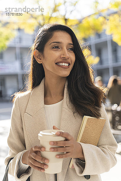 Lächelnde junge Frau mit Buch und Kaffee zum Mitnehmen in der Stadt  die sich umsieht