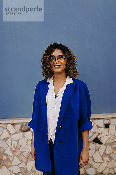 Porträt einer lächelnden Frau mit Brille und blauem Blazer  blaue Wand im Hintergrund