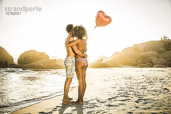 Paar mit herzförmigem Ballon  das sich bei Sonnenuntergang am Strand umarmt