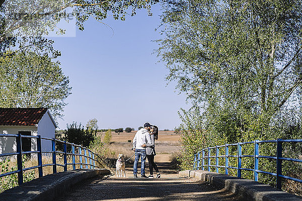 Ein küssendes Paar mit seinem Hund  das auf einer Brücke in einem Park steht