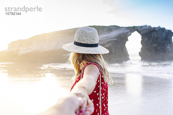 Blonde Frau in rotem Kleid und Hut  die am Strand Hand hält  Naturbogen an der Playa de Las Catedrales  Spanien