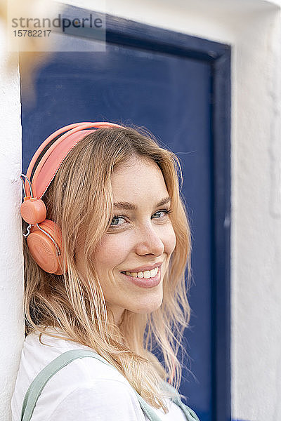 Porträt einer glücklichen jungen Frau  die sich an die Fassade lehnt und mit Kopfhörern Musik hört