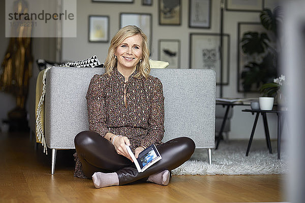 Porträt einer lächelnden blonden Frau  die zu Hause auf dem Boden sitzend entspannt