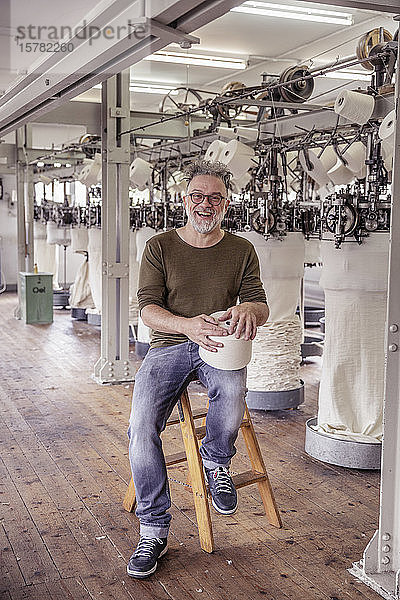 Porträt eines glücklichen reifen Mannes  der auf einer Leiter in einer Textilfabrik sitzt