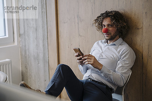 Porträt eines Geschäftsmannes mit roter Clownsnase bei der Benutzung eines Mobiltelefons im Büro