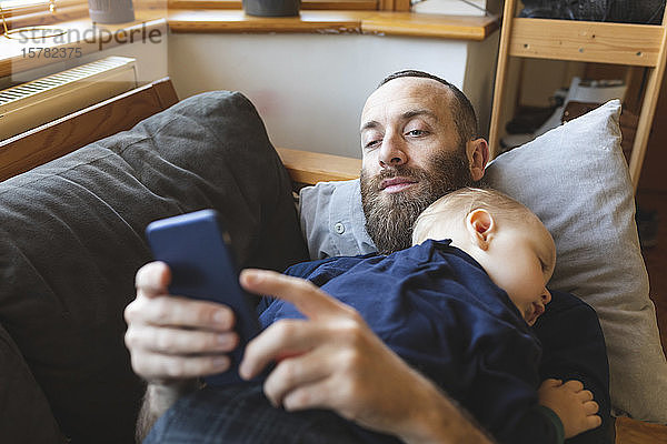 Mann überprüft sein Telefon  während sein kleiner Sohn auf dem Sofa schläft
