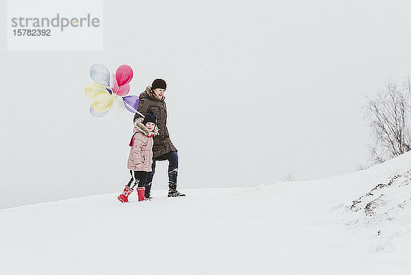 Mutter und kleines Mädchen mit Luftballons gehen in verschneiter Landschaft