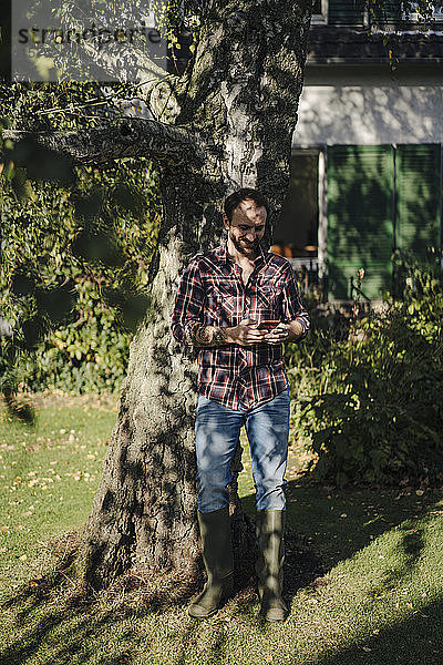 Reifer Mann mit Gummistiefeln unter einem Baum stehend  mit Smartphone