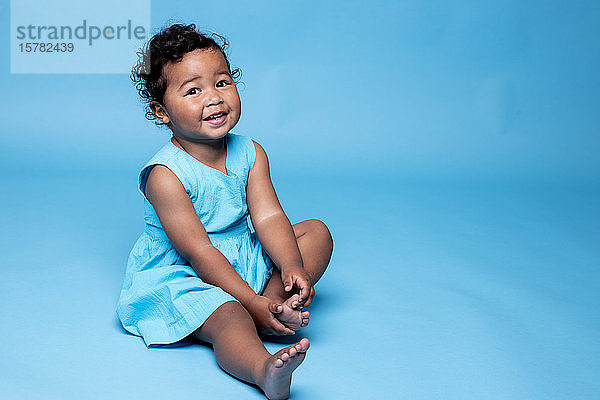 Bildnis eines barfüssigen kleinen Mädchens in hellblauem Kleid vor blauem Hintergrund sitzend