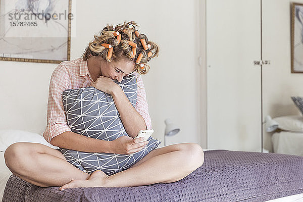 Glückliche junge Frau mit Lockenwicklern im Haar sitzt auf dem Bett und schaut auf ihr Handy