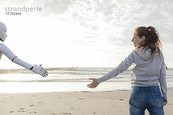 Lächelnde Frau steht am Strand und greift nach Roboterhand