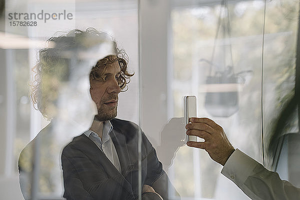Geschäftsmann zeigt seinem Kollegen hinter einer Glasscheibe sein Handy