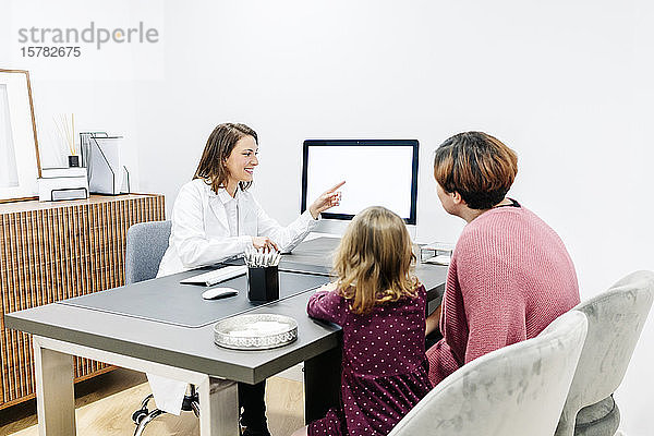Arzt im Gespräch mit Mutter und Tochter am Schreibtisch in der Arztpraxis