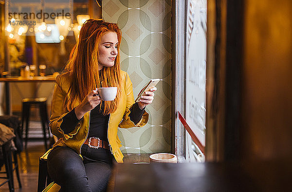Rothaarige junge Frau sitzt in einem Cafe mit einer Tasse Kaffee und schaut auf ihr Handy