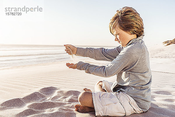Kleiner Junge sitzt am windigen Strand und beobachtet Sand  der aus seinen Händen rieselt