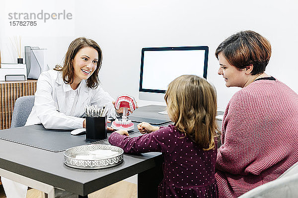 Zahnarzt zeigt Mutter mit Tochter Zahnmodell am Schreibtisch in medizinischer Praxis