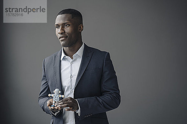 Porträt eines jungen Geschäftsmannes mit Spielzeugroboter vor grauem Hintergrund