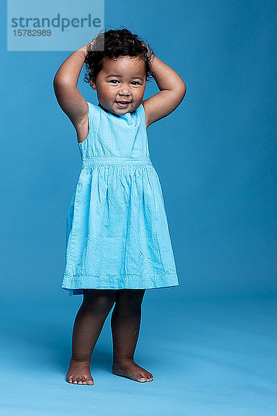 Bildnis eines lächelnden kleinen Mädchens mit Händen auf dem Kopf in hellblauem Kleid vor blauem Hintergrund stehend