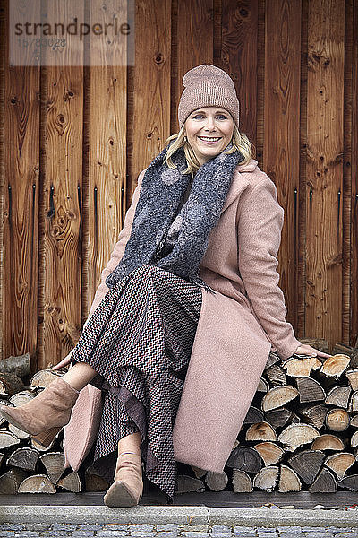 Bildnis einer lächelnden Frau in Winterkleidung auf einem Holzstapel sitzend