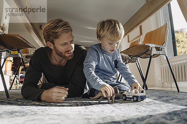 Vater und Sohn spielen zu Hause mit Dinosaurierfigur und Spielzeugauto unter dem Tisch