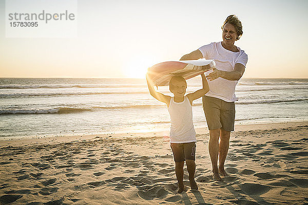 Glückliche Vater und Sohn mit Surfbrett bei Sonnenuntergang am Strand spazieren