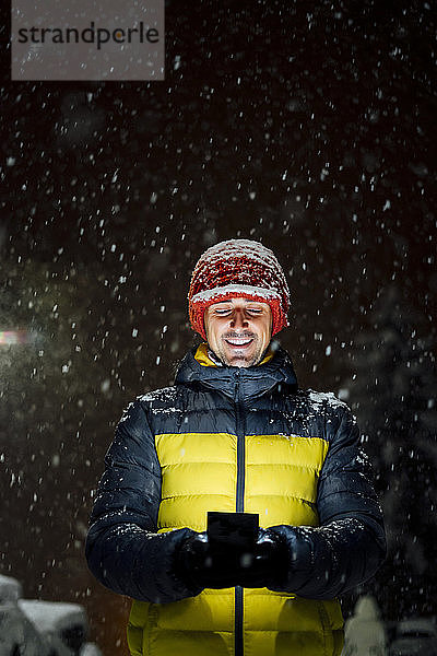 Lächelnder Mann benutzt Mobiltelefon bei Schneefall in der Nacht