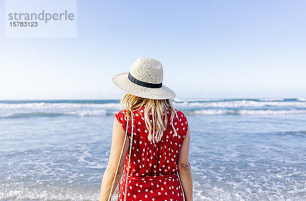 Blonde Frau in rotem Kleid und Hut  die am Strand spazieren geht  Playa de Las Catedrales  Spanien