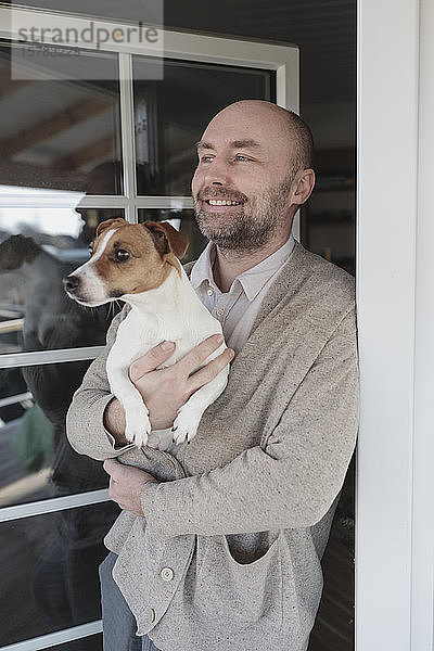 Porträt eines entspannten Mannes mit Hund auf den Armen  der an der offenen Terrassentür seines Hauses steht