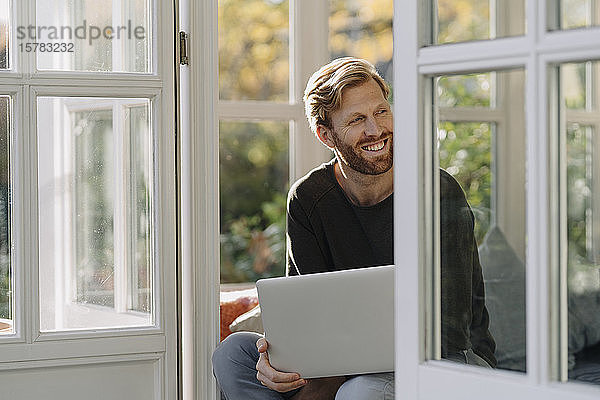 Lächelnder Mann mit Laptop im Wintergarten zu Hause