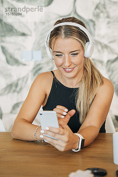 Lächelnde Frau am Tisch sitzend mit Kopfhörern und Smartphone