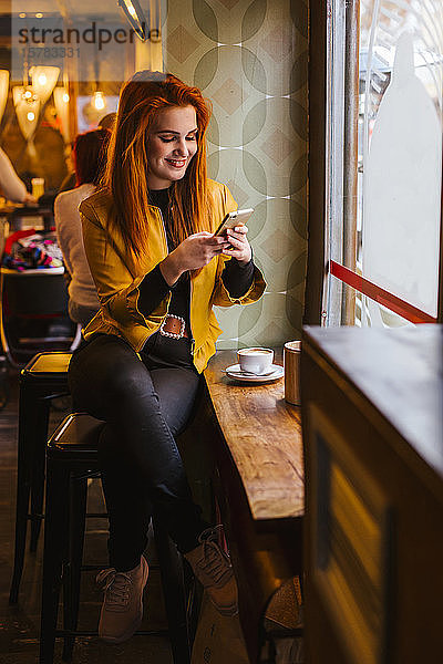 Porträt einer glücklichen rothaarigen jungen Frau  die mit ihrem Handy in einem Café sitzt