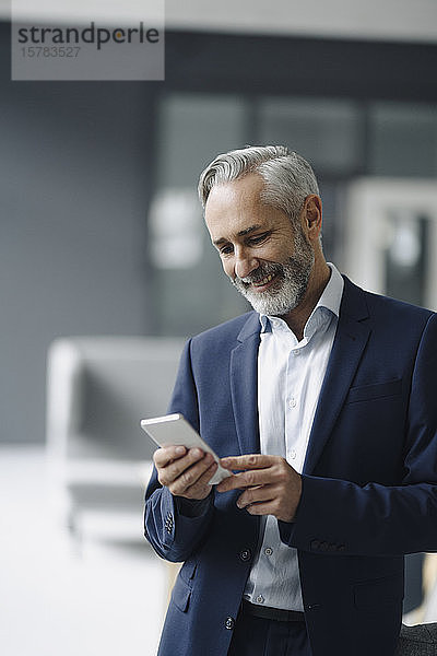 Porträt eines lächelnden reifen Geschäftsmannes in seinem Büro  der auf ein Smartphone schaut