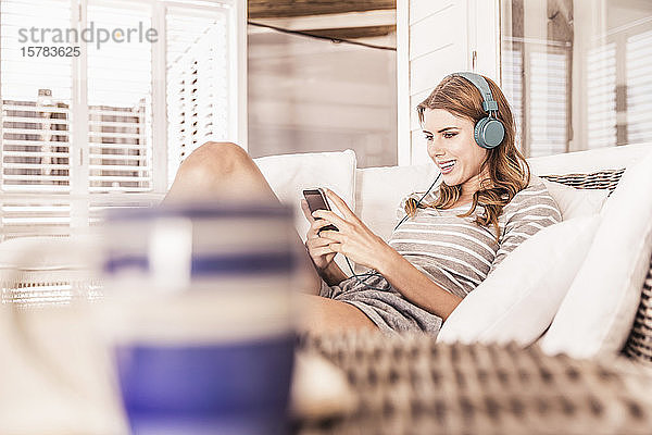 Junge Frau entspannt sich auf einer Veranda mit Smartphone und Kopfhörern