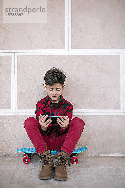 Junge sitzt mit einem Smartphone auf einem Skateboard an einer Wand