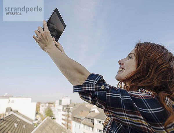 Lächelnde rothaarige Frau mit Tablette auf der Dachterrasse beim Selfie
