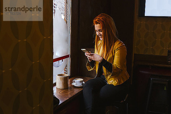Glückliche rothaarige junge Frau sitzt in einem Café und benutzt ihr Handy