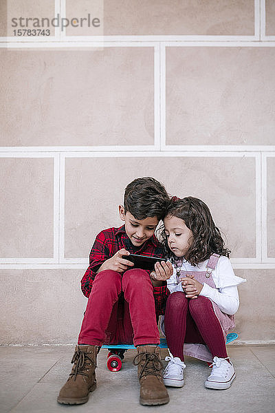 Junge und Mädchen sitzen mit einem Smartphone auf einem Skateboard an einer Wand