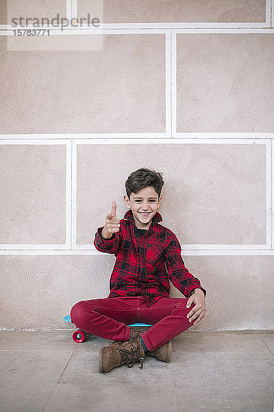 Porträt eines glücklichen Jungen  der auf einem Skateboard an einer Wand sitzt