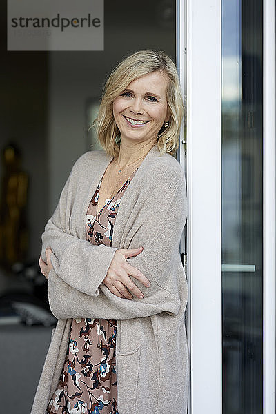 Porträt einer lächelnden blonden Frau  die sich zu Hause an die Balkontür lehnt