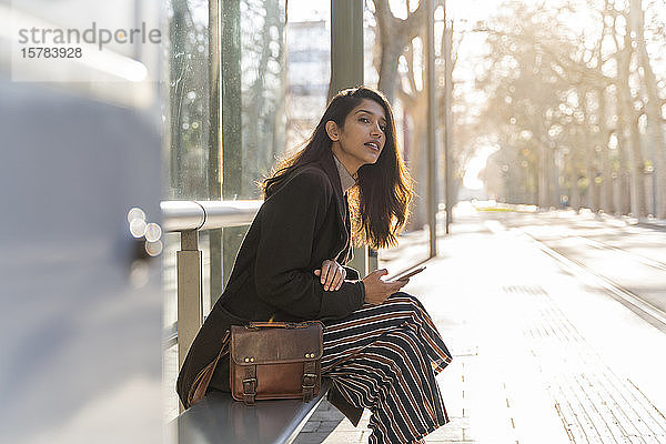 Junge Frau mit Smartphone wartet an der Straßenbahnhaltestelle