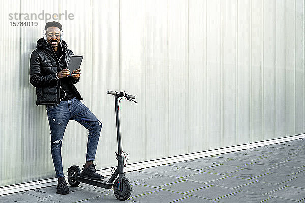 Porträt eines glücklichen Mannes mit Kopfhörer  Tablet und E-Scooter im Freien