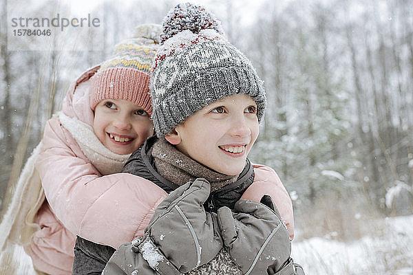 Porträt eines Jungen  der seine kleine Schwester im Winterwald Huckepack reitet