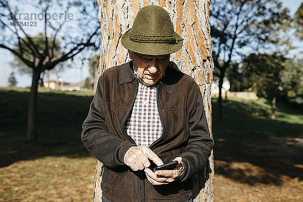 Alter Mann mit Smartphone  an Baumstamm gelehnt