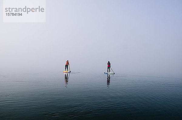 Zwei Frauen stehen beim Paddelsurfen auf einem See im Nebel auf