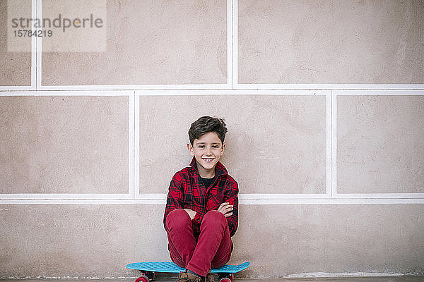 Porträt eines lächelnden Jungen  der auf einem Skateboard an einer Wand sitzt