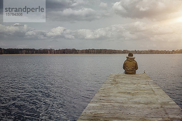 Rückenansicht eines jungen Mannes  der auf einem Steg sitzt und im Winter auf den See schaut