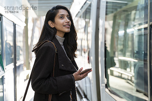 Lächelnde junge Frau mit Smartphone an der Straßenbahnhaltestelle