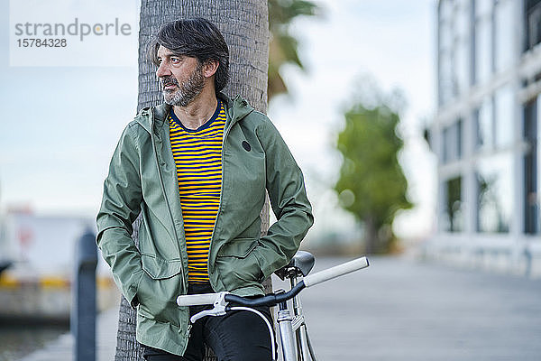 Porträt eines seriösen reifen Mannes mit einem Fahrrad  das an einen Palmenstamm gelehnt ist und in die Ferne schaut  Alicante  Spanien