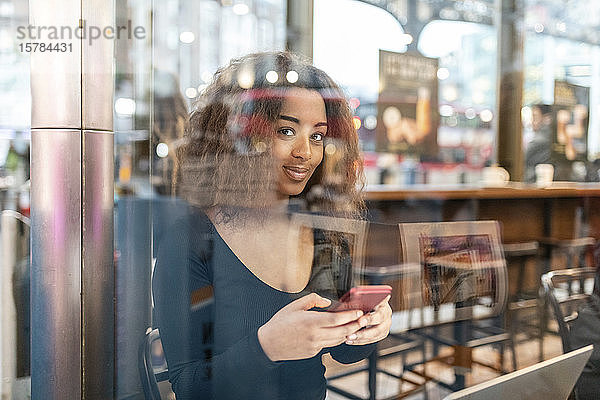 Porträt einer lächelnden jungen Frau  die ein Smartphone in einem Cafe benutzt
