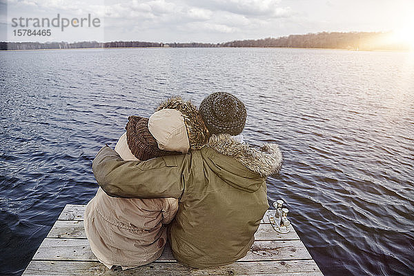 Rückenansicht eines jungen verliebten Paares  das auf einem Steg sitzt und im Winter auf den See schaut